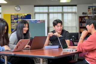 Group photo of four college students talking to each other while sitting at a table and working on their laptop computers. 