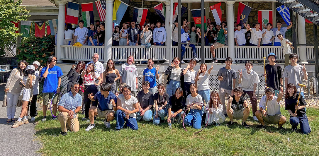 A group photo of about 60 international students standing on the porch and the lawn in front of a historic academic building decorated with colorful national flags.