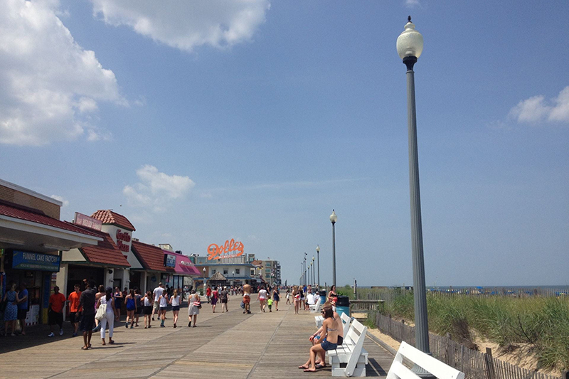People walk along a beach boardwalk lined with stores, white benches and light posts.