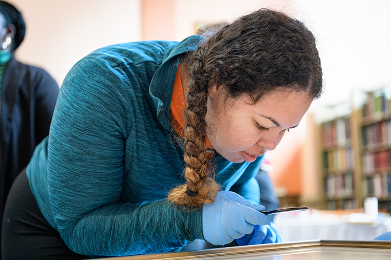 A graduate student wearing blue latex gloves uses a magnifying glass to inspect an item at the Delaware Historical Society Research Library in Wilmington, Delaware.