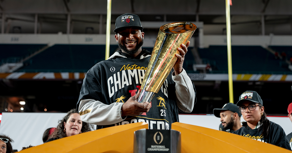 UD alumnus Isaac Hicks, director of sports performance nutrition for Indiana University Bloomington, holds the National Championship Trophy, which is gold and oblong-shaped like a football at the base, tapering up to a flattened full-size football at the top.