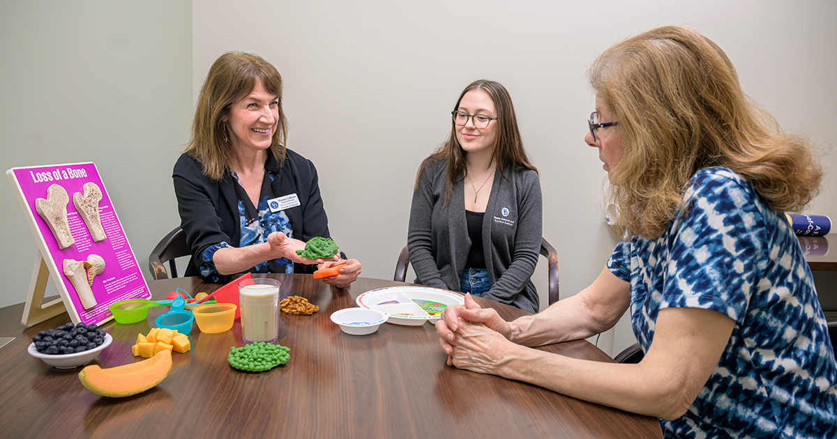 Sharon Collison, registered dietitian in UD's Nutrition Clinic, holds plastic fruit and vegetable models while sitting around a table with a sudent and a client.