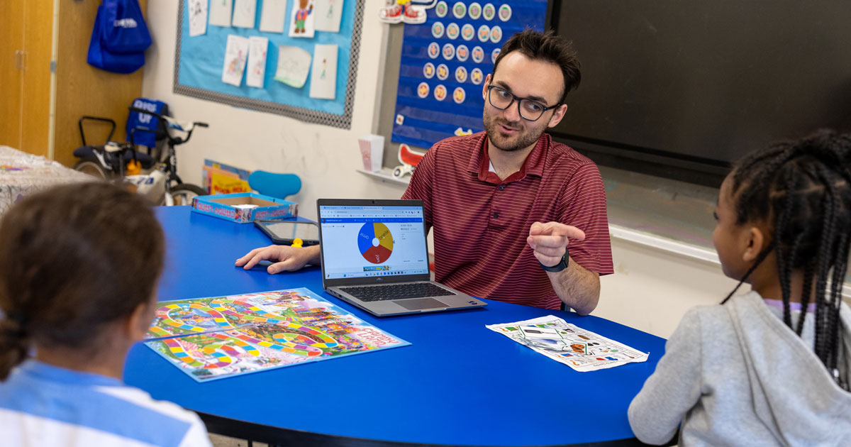 Jon Tressler, a 2024 graduate from the Speech-Language Pathology master’s program in UD’s College of Health Sciences, works with kids at Brader Elementary School. Pictured here, Mr. Jon, as students call him, has students practicing pronouncing “L” words and describing objects in complete sentences.  He's seen pointing vat the students while showing them a laptop screen on a bright blue table.