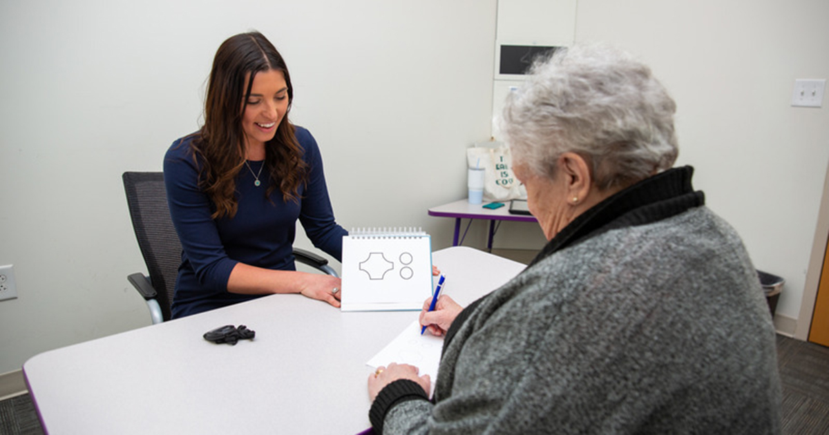 Alyssa Lanzi, assistant professor of communication sciences and disorders, sits across the table from a woman and conducts a cognitive test. On a notepad facing the test subject are shapes. The test subject is holding a pen. 