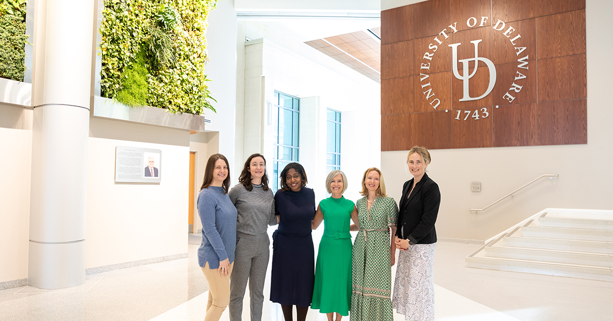 Winston Churchill Fellow Heather McFarlane (middle, wearing a green dress), stands with Freda Patterson, associate dean of research for CHS, and Epidemiology and Partnership for Healthy Communities faculty and staff in the lobby of STAR with a large UD logo behind them.