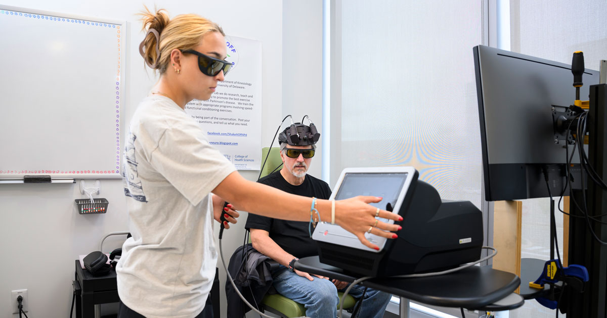 Interdisciplinary neuroscience Ph.D. student Sara Penuela Rodriguez is wearing goggles as she administers infrared laser therapy through a touchscreen computer to study participant Tim Hihn who's shown in the background wearing a helmet iwth sensors eqipped to it to the deliver the light. The study is blind and it's unclear whether Hihn is receiving the laser therapy or a placebo.