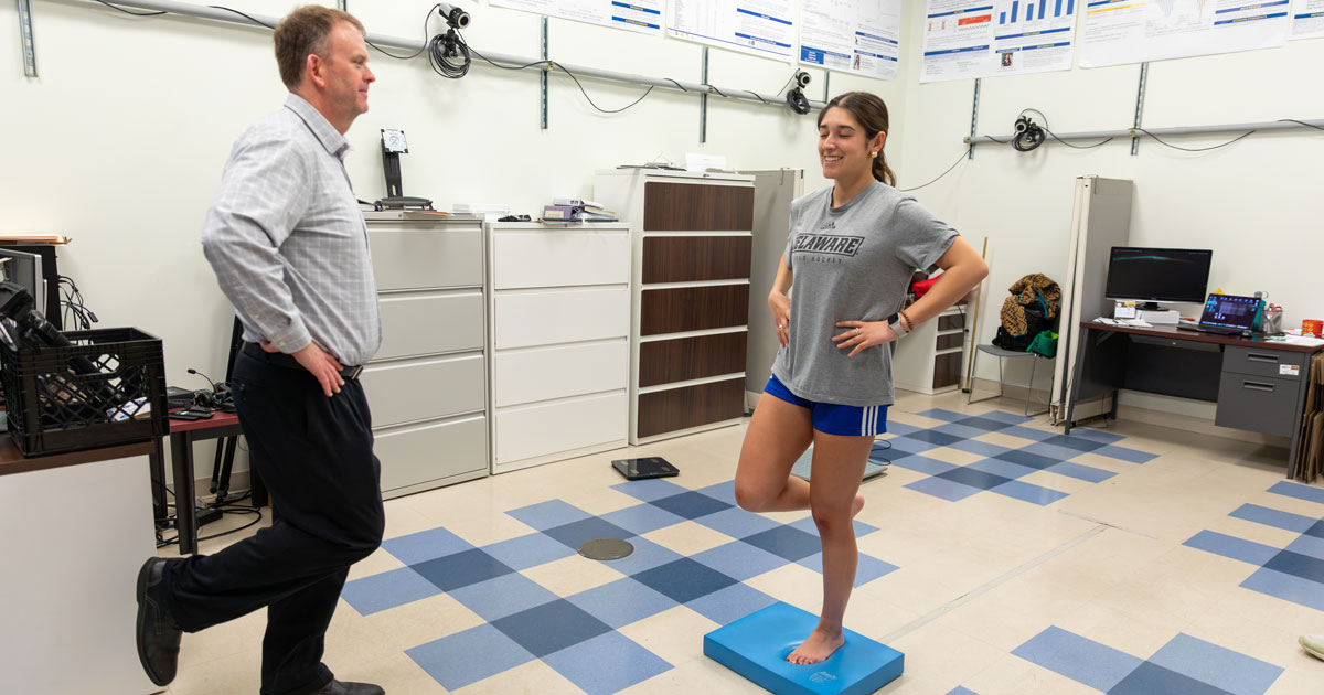 UD field hockey player Izzy Rothwell, stands on one foot on a mat while Tom Buckley, professor of kinesiology and applied physiology, also stands on one foot facing her, demonstrating baseline concussion testing.