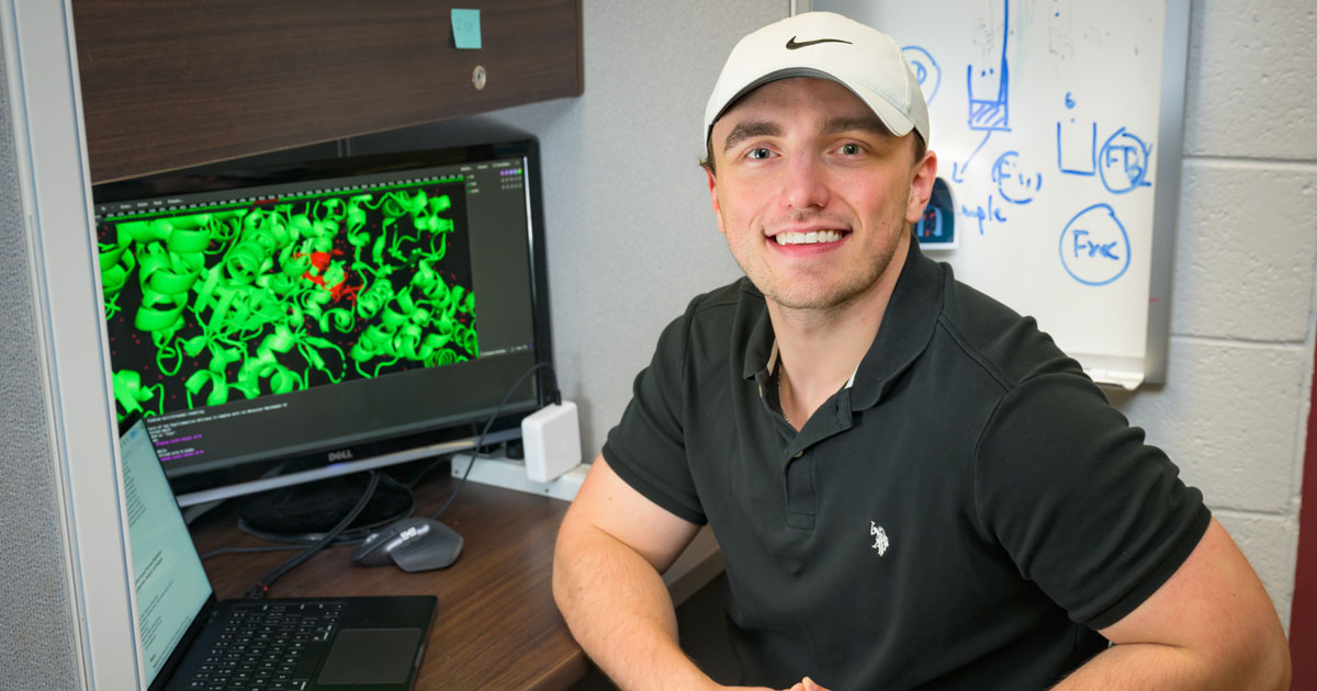 Sean Fletcher, a senior honors medical diagnostics student, sits at a desk facing the camera and to his side is a computer screen showing green HPV virus molecules. He's wearing a white Nike baseball caap and a black shortsleeve polo.