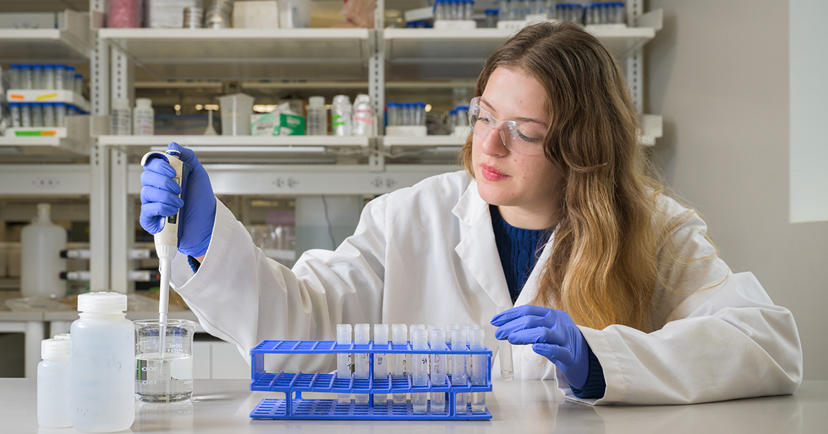Corinne Van Hoven, a senior honors medical diagnostics major, works with a laboratory sample holding a pipette, putting a liquid into a glass jar. She's wearing a white lab coat, goggles and blue latex gloves and has other laboratory equipment around her and behind her.