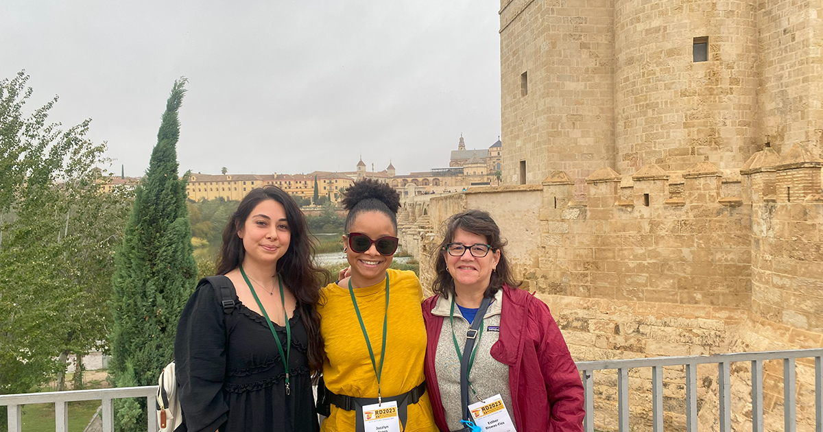 From left to right, medical sciences doctoral students Senem Cevik and Jazzlyn Jones pose with Medical and Molecular Sciences Professor and Chair Esther Biswas-Fiss with an ancient castle or fortress-like structure in the background in Spain