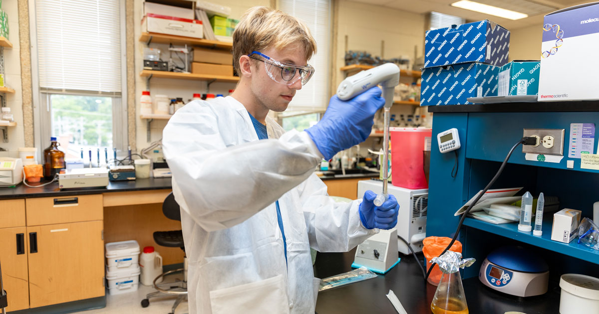 John Routzahn, who graduated from UD in 2024 with a bachelor’s degree in applied molecular biology and biotechnology, is shown here working in a UD lab. He's wearing a white lab coat, goggles, and blue gloves while handling a specimen.