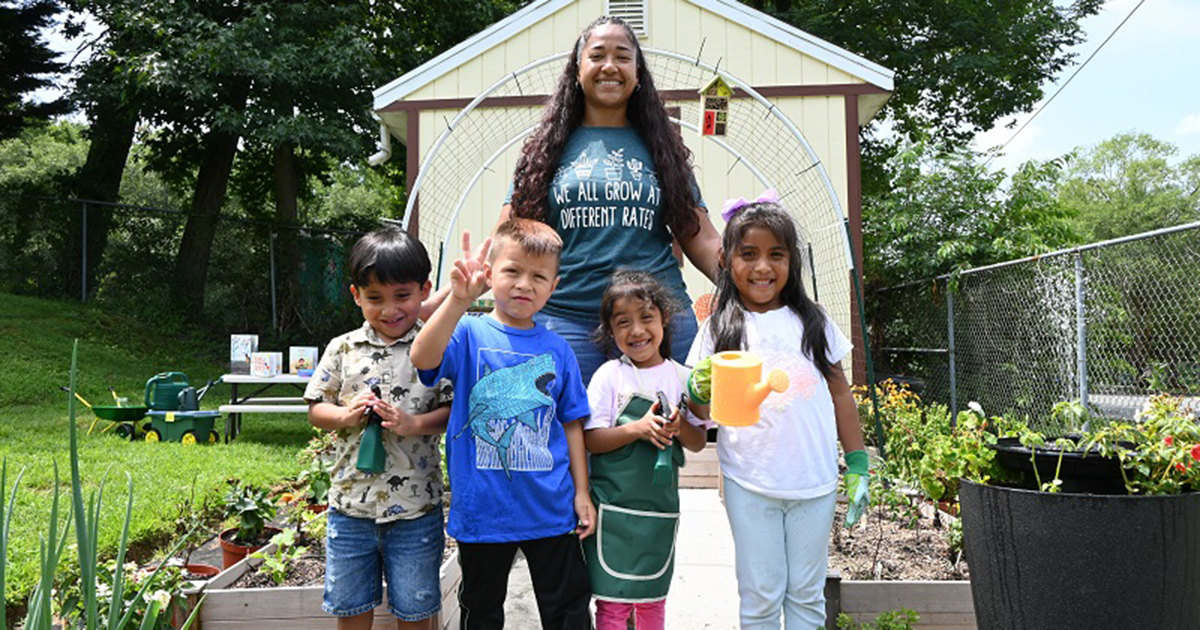 Diana Mercado, early childhood administrator at Pirulo’s Child Care and Learning Center, smiles with four children in front of the garden she grew with the support of UD’s Let’s Grow Outside! initiative. One child is giving a peace sign. One is holding a yellow watering can. The other two children are holding gardening tools. 