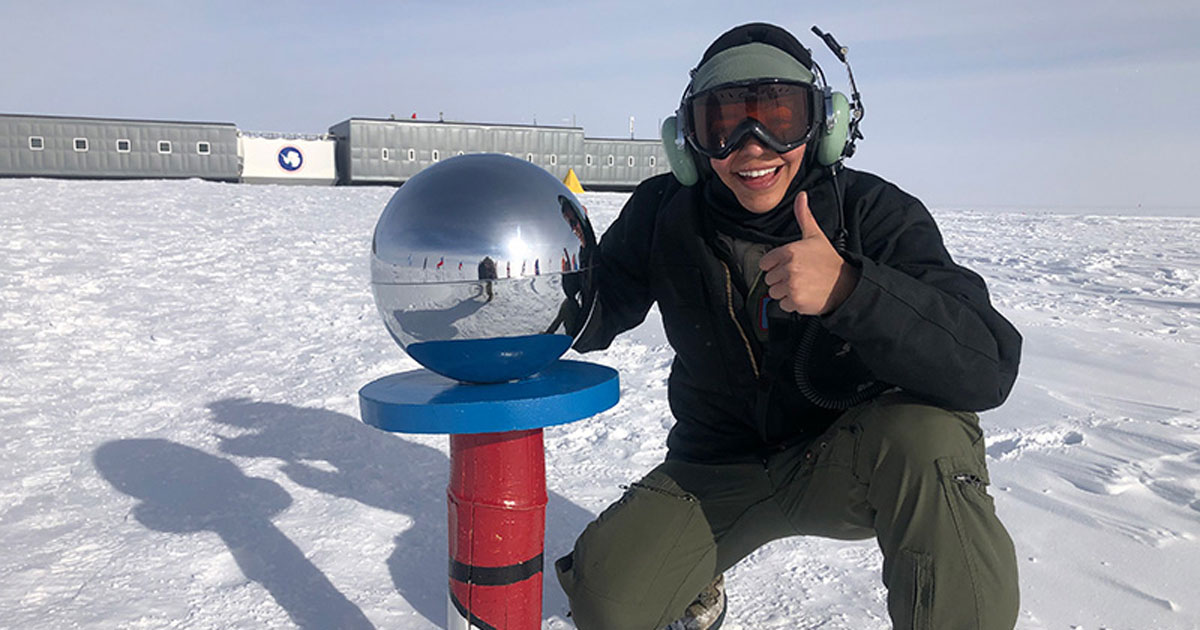 Katy Buoncristiano, a second-year speech-language pathology master's student, poses at the bottom of the world at the ceremonial marker for the South Pole giving a thumbs up sign. She's wearing a full snow suit and goggles and a headset. Snow is behind her along with the iconic Amundsen-Scott South Pole Station.