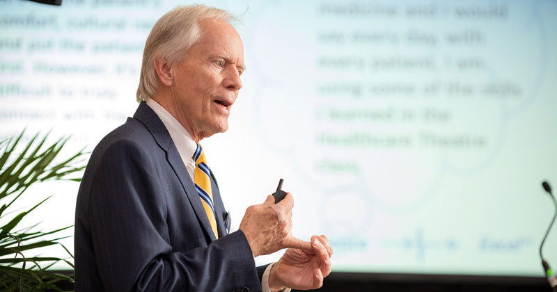 Allan Carlsen discusses Healthcare Theatre at a University event in Baltimore in 2018. A white screen with blurred text is the backdrop.
