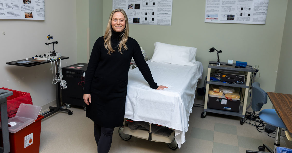 Megan Wenner, associate professor of kinesiology and applied physiology wears a black dress and black tights with her hand leaning on a research bed in a lab. Laboratory items are in the background. 