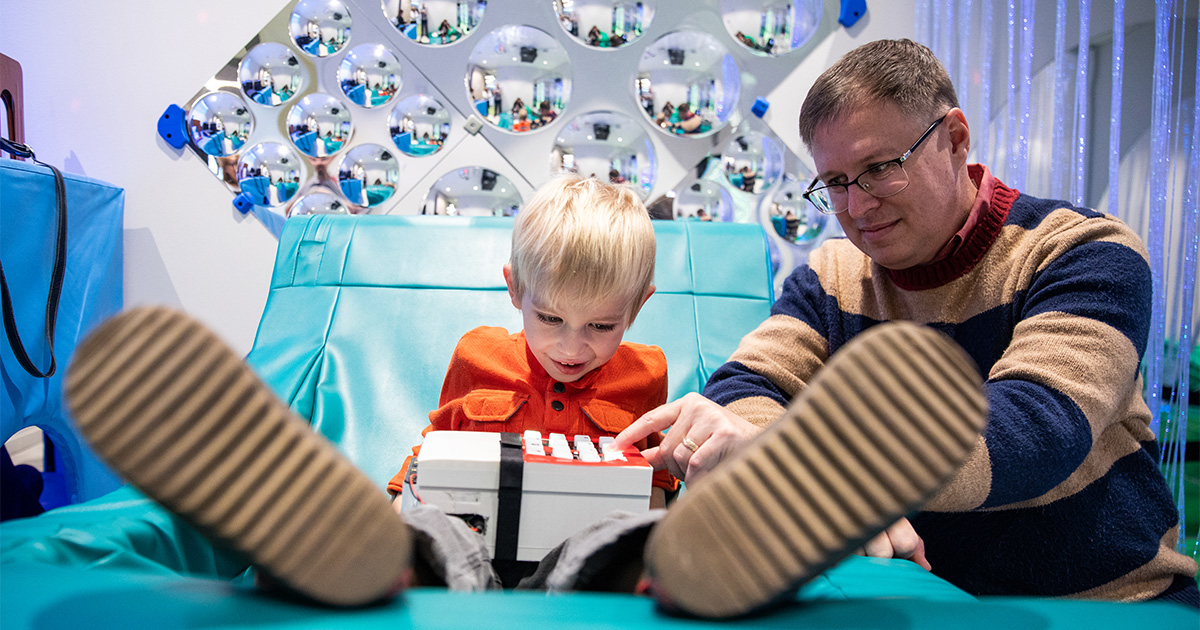 A young boy in an orange shirt sits in a teal-colored chair in the Route 9 Library and Innovation Center's colorful Sensory Room. Here, he's learning how to use a new device that helps children with autism modify music to suit their listening needs. In this photo, his father shows him how to use the device.