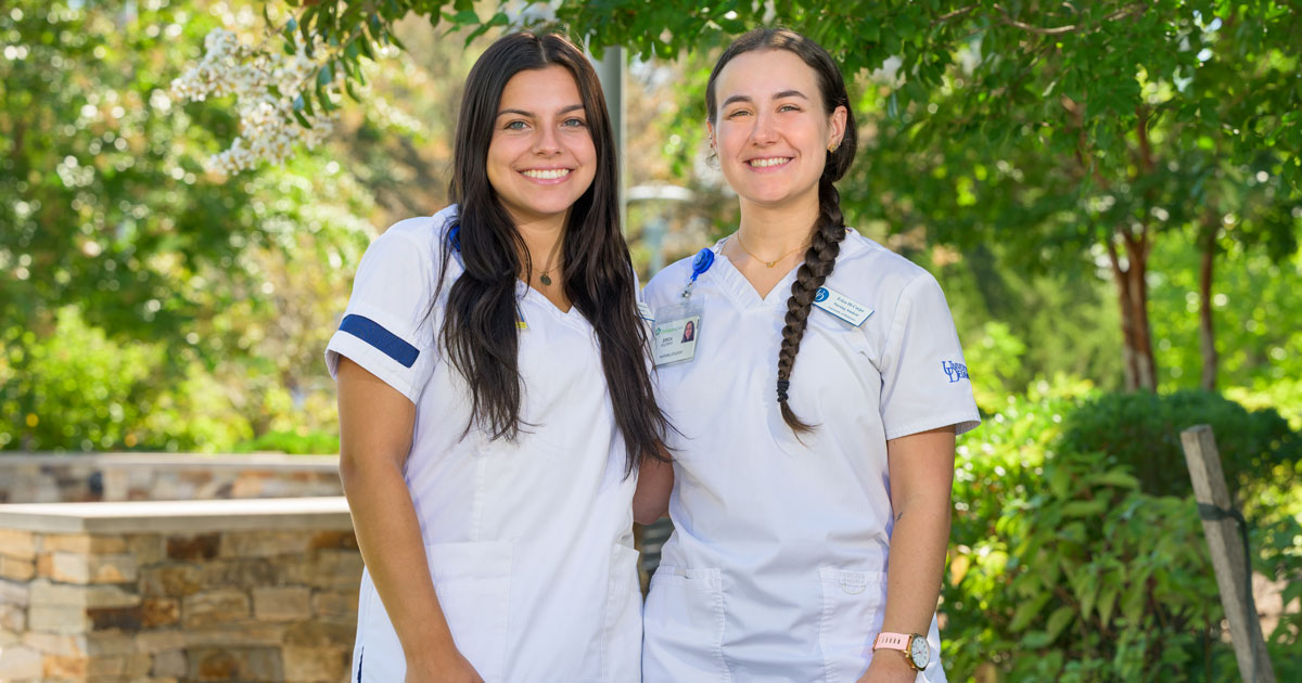 Two School of Nursing seniors stand side by side in their white and blue UD nursing scrubs surrounded by greenery on UD's campus.