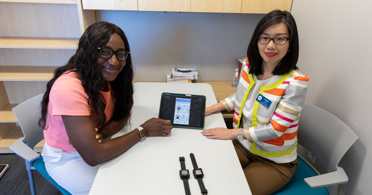Fatou Marong, a master’s student in the health promotion program, (left) and Xiaopeng Ji, associate professor of nursing (right), are seated at a rectangular table with an ipad demonstrating a sleepchat bot they created. Also on the table are two fitness bands that help monitor sleep.