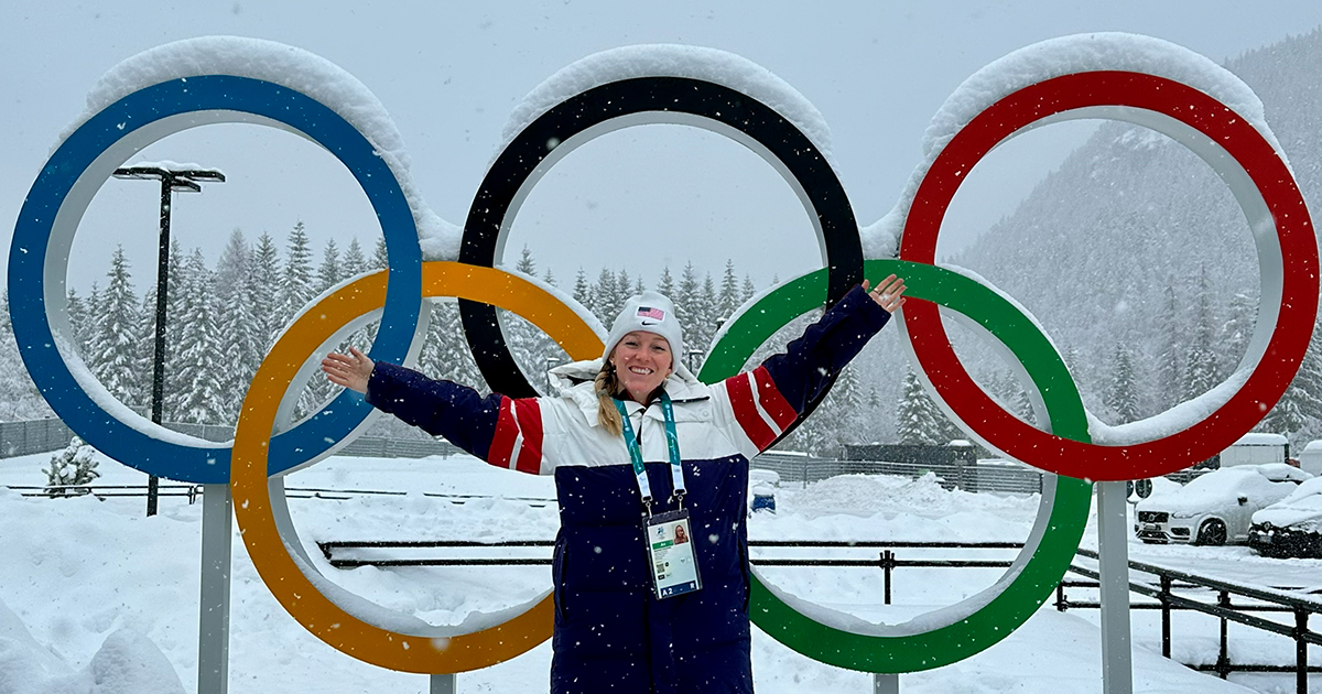 Shannon Colleton, a UDPT sports residency alumna and now sports physical therapist with U.S. Ski and Snowboard,  wears her official red white and blue long ski jacket, a white hat with the American flag on it, and a lanyard with her credentials on it. She's standing in front of the iconic multi-colored Olympic rings with her arms in the air with snow falling around her.