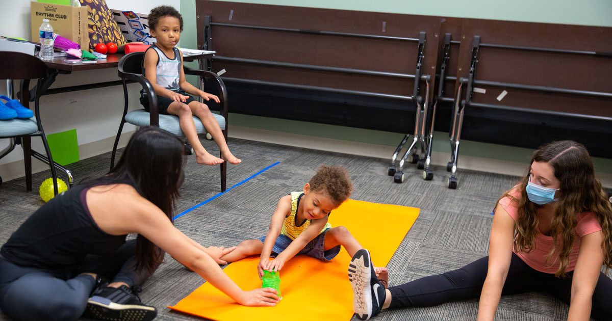 A child plays with a green cup using whole-body movements while seated on an orange mat with two research assistants sitting on the floor. Another child sits in a chair watching his brother.