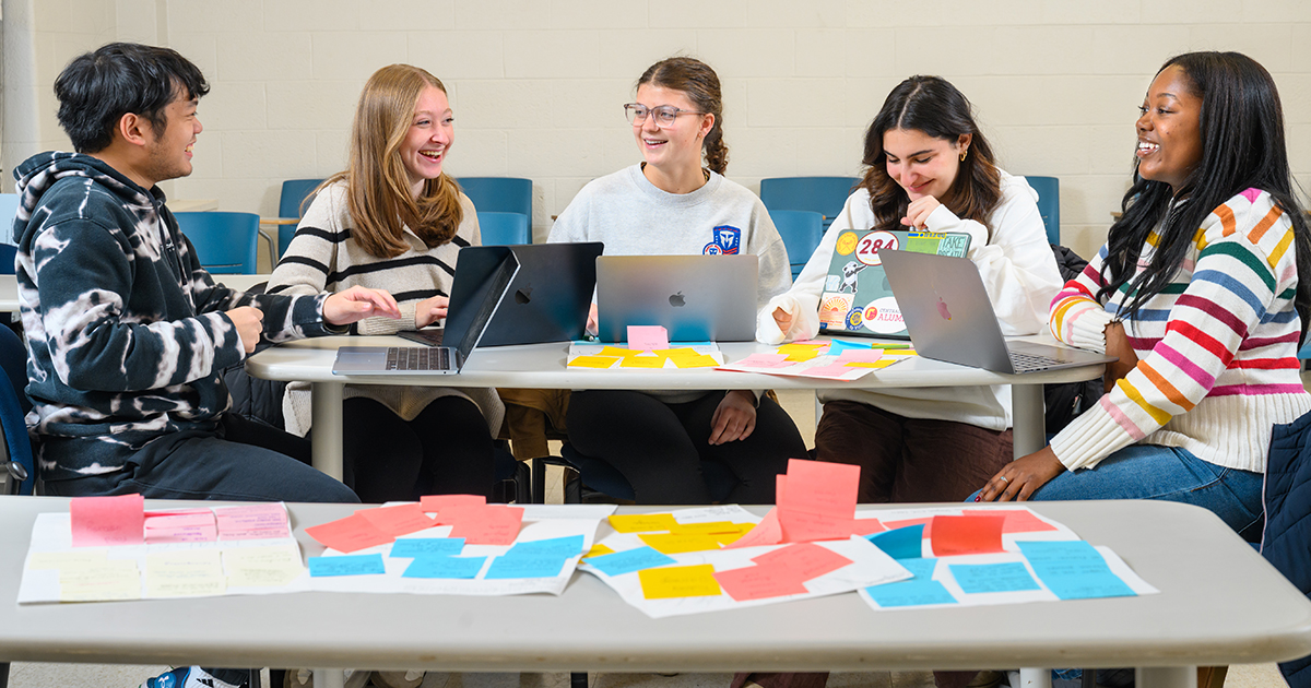 First-year nursing students sit around a table with laptops working together. In front of them are multiple pink, blue, and orang sticky notes representing a concepts and ideas to show a problem tree.