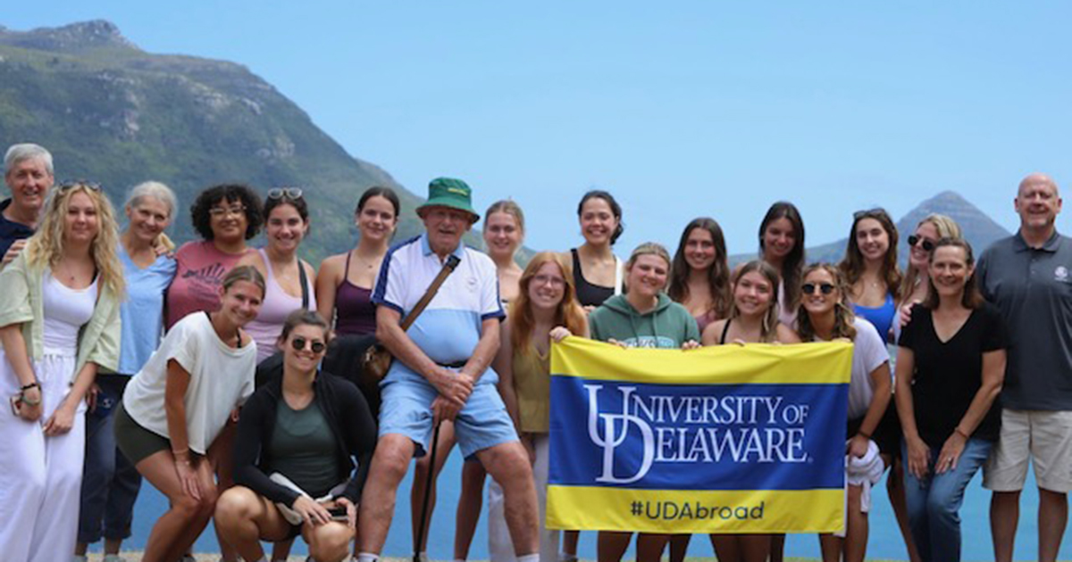 Students and trip leaders stop along the scenic Chapman’s Peak Drive in Cape Town to admire the view.  They're holding the blue and yellow UD flag with mountains and blue sky behind them.