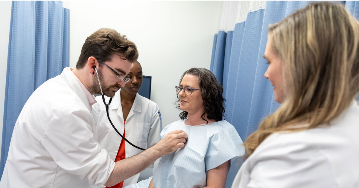 A nurse practitioner student uses a stethoscope to listen to a patient's heartbeat while another student and Amanda Watson, assistant professor of nursing, watches.