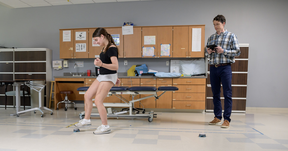 Peyton Harter hops and steps between buzzers as part of her recovery in UD's Physical Therapy Clinic while PT Jimmy Orr looks on holding an iPad-like device. 