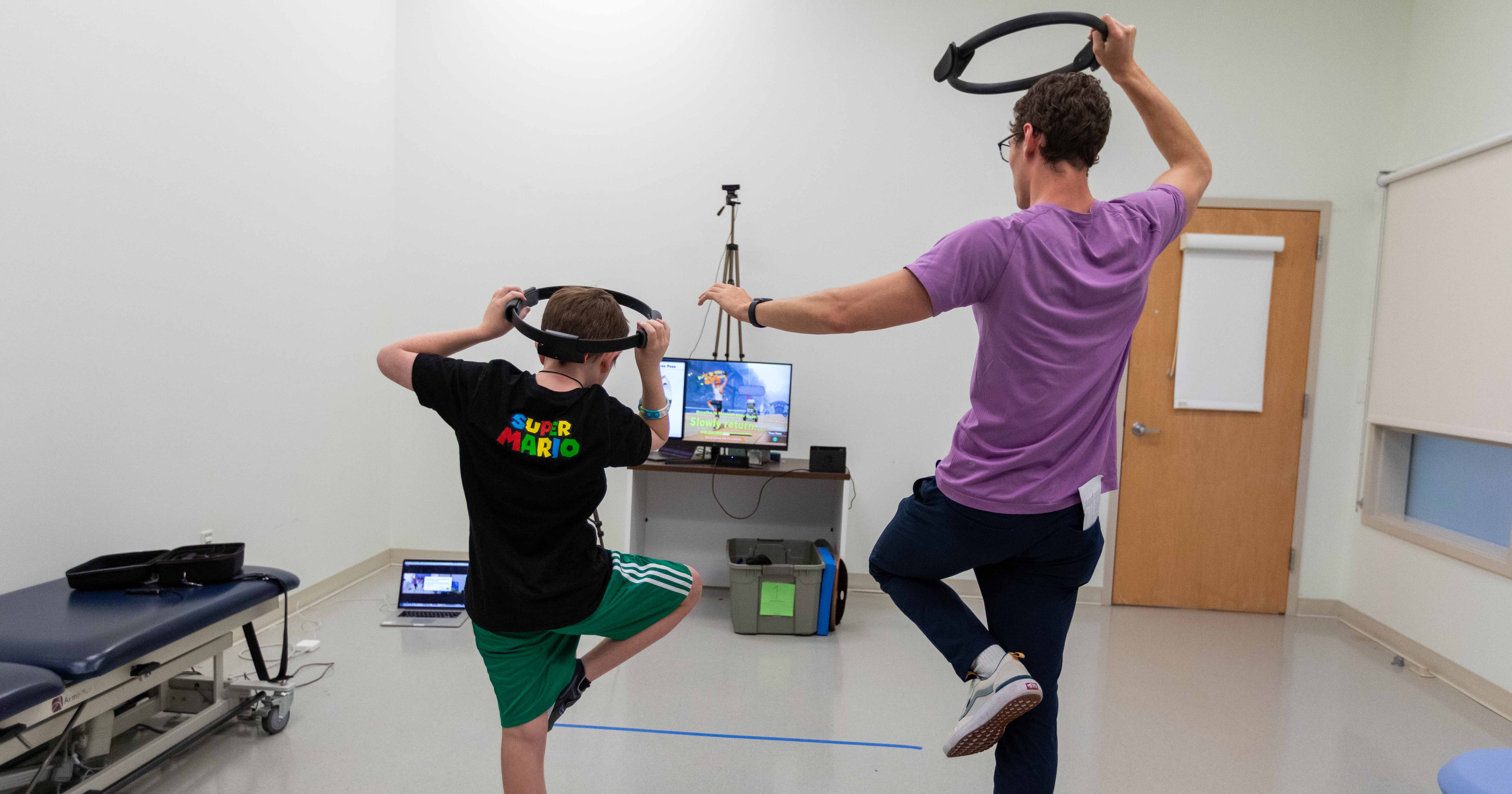 A 10-year-old boy with autism and Jacob Corey (right), a research assistant obtaining his doctorate in biomechanics and movement science, play with Ring Fit. In this photo, their backs are turned to the camera, while they watch a video game screen, and each hold a ring over their heads while standing on one leg. They're testing Ring Fit as a gross motor intervention for children with autism. 