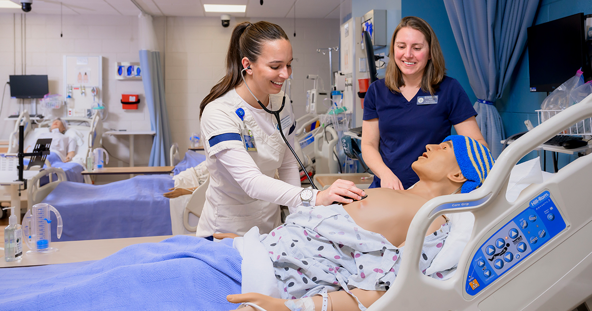 Senior nursing major Madison Wirchansky (left), wearing her white and blue UD nursing uniform, uses a sthetoscope to listen to heartbeat and breath sounds on a high-fidelity manikin lying in a bed while Michelle DePhillips, assistant professor of nursing, wears navy scrubs and looks on in UD's Simulation Lab. 