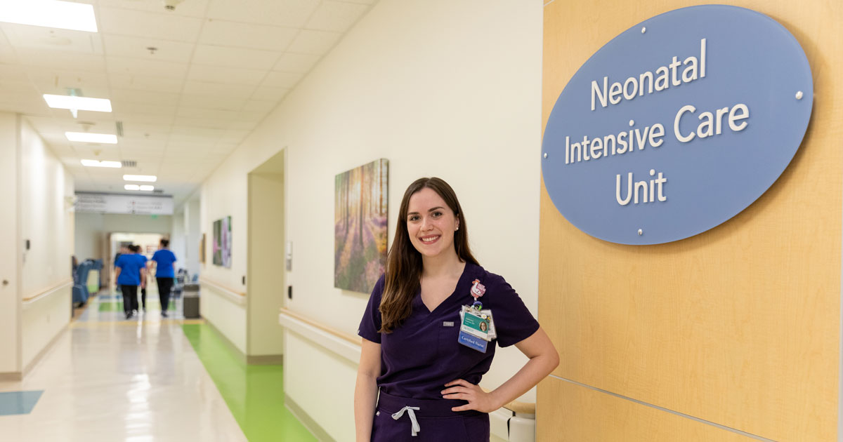UD alumna Cat Lawless stands in her purple scrubs outside the periwinkle oval-shaped Neonatal Intensive Care Unit sign at Nemours Children's Hospital where she works. The sign is on a yellow wall, and there's a long hallway behind Lawless, who's wearing purple scrubs and has one hand on her hip.