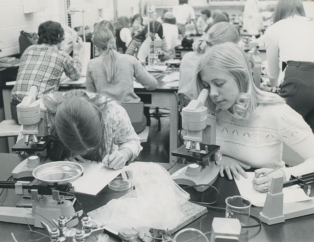 A historic image of two female students in a lab looking through a microscope