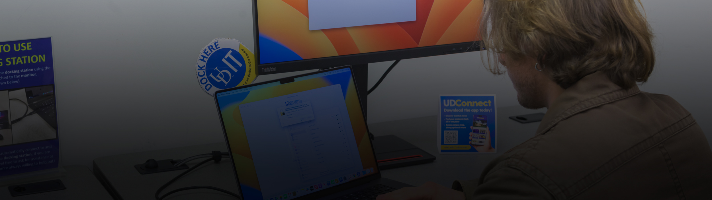 A student works on a laptop connected to an external monitor, with a "Dock Here" sticker and "UDConnect" sign visible on the desk.