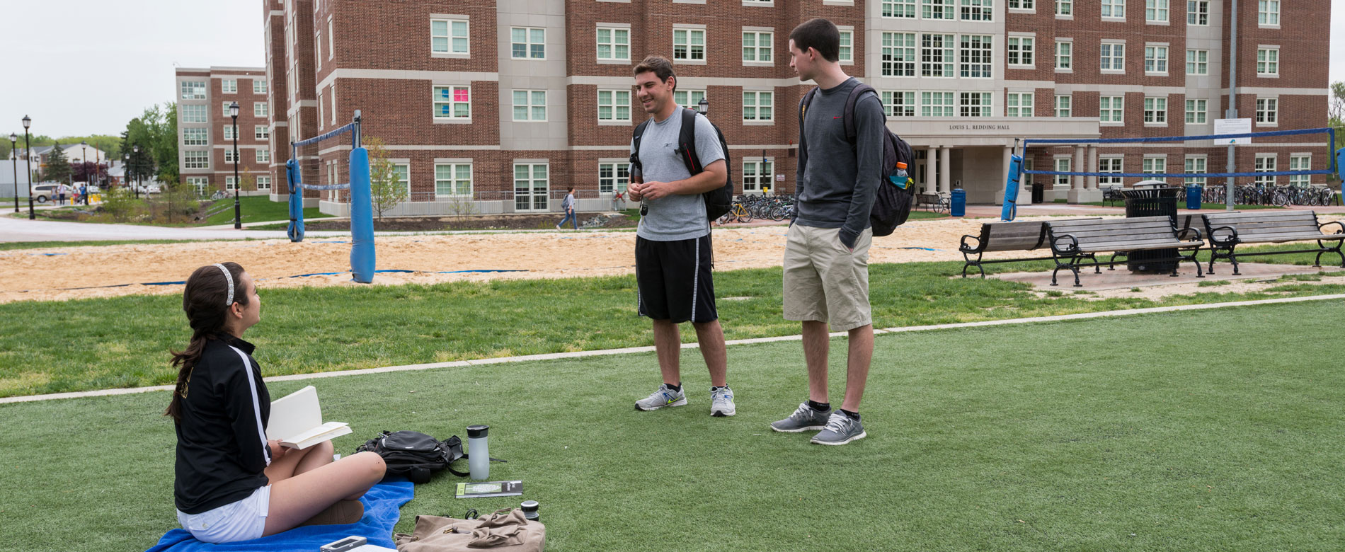 Students stand on the Turf talking; a volleyball court in the background