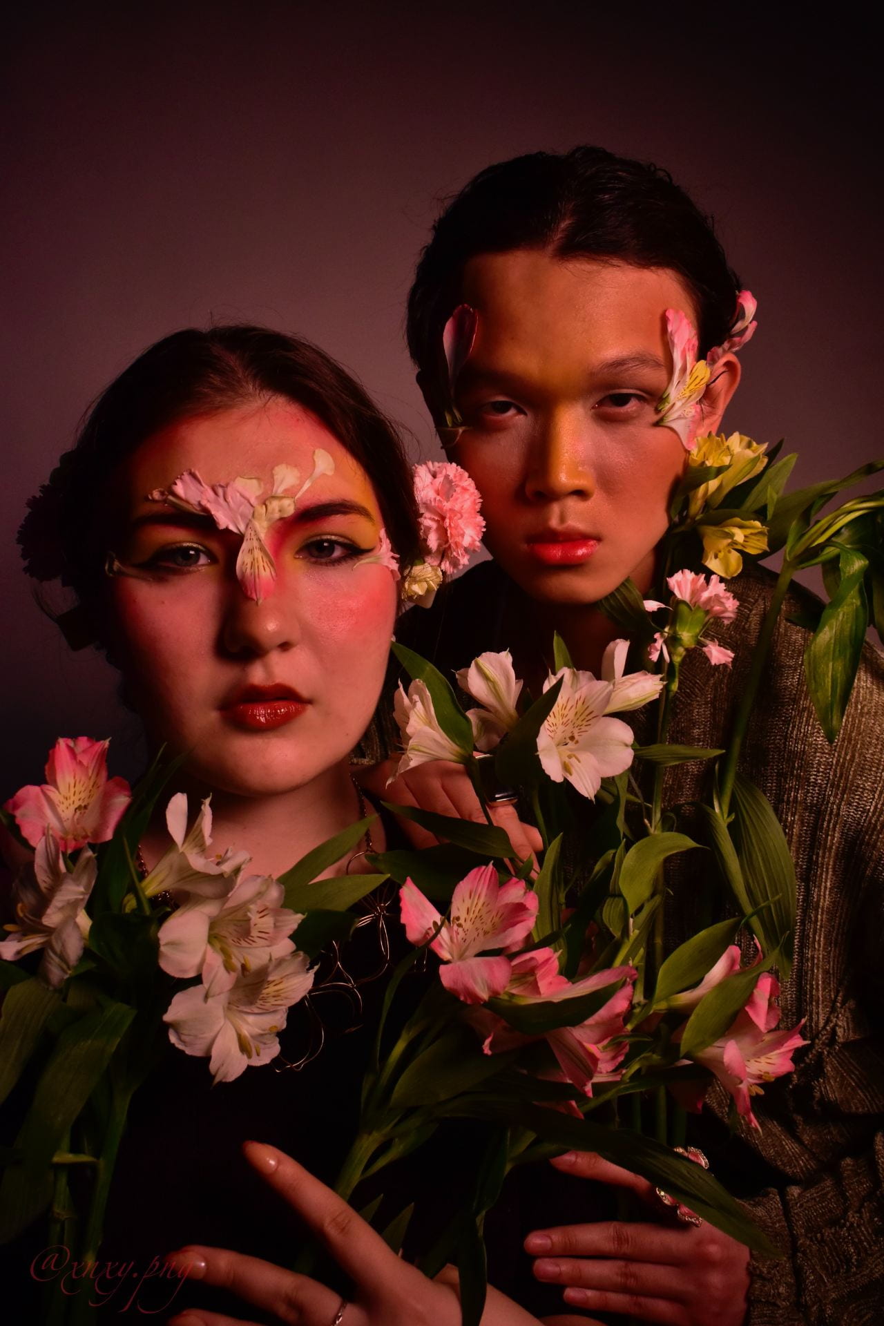 Photograph of two people cast in a pink light. They are surrounded and adorned by pink flowers