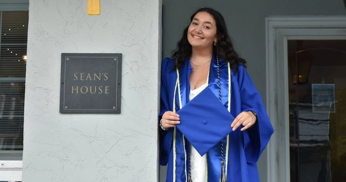 Missy Marazzo in UD blue cap and gown stands on porch of Sean's House