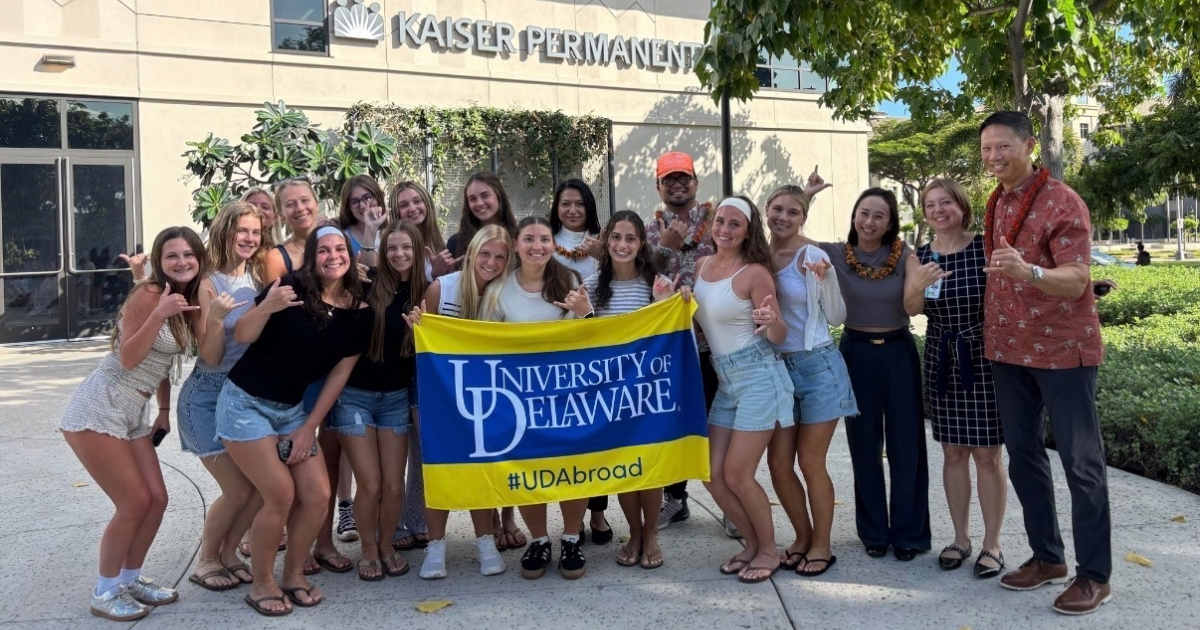 Group of UD students hold UD Study Abroad blue and gold flag in front of Kaiser Permanente in Hawai'i