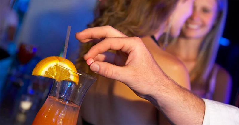 A man drops pill into an orange drink while women stand in the background