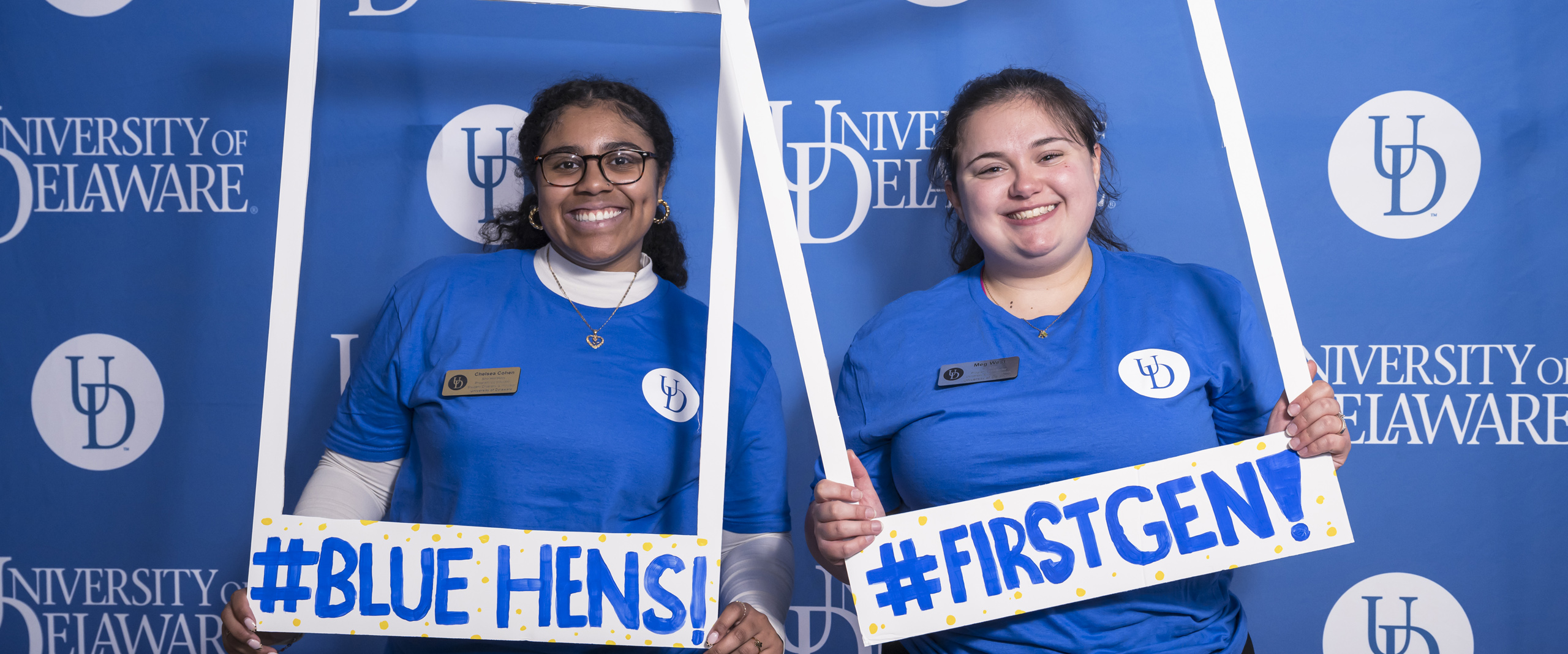 two staff members pose with Polaroid-style frames reading "#BLUEHENS!" and "#FIRSTGEN!"