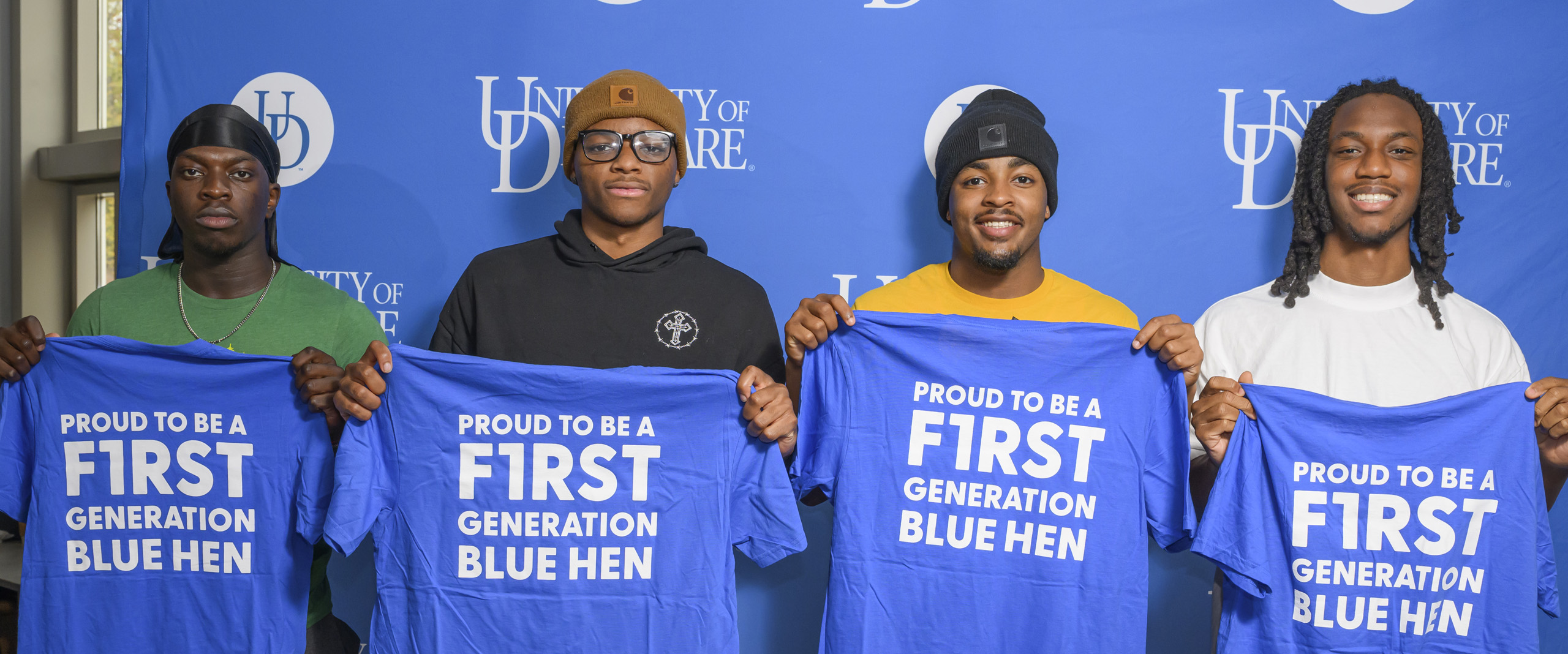 4 first-generation students pose holding blue t-shirts reading "PROUD TO BE A F1RST GENERATION BLUE HEN"