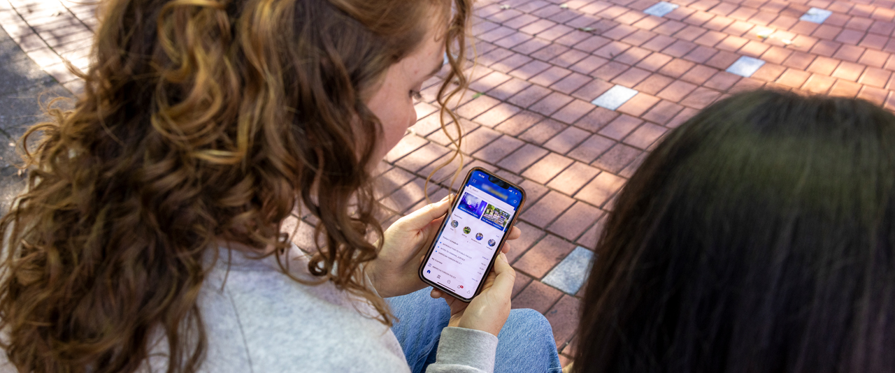 two students sitting on a bench look at a UD web page on a mobile phone