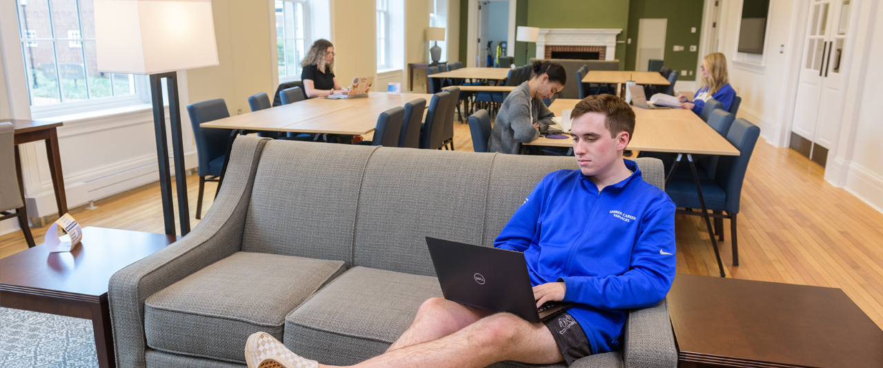 A student sits on a couch and works on a laptop, while other students work at tables behind him