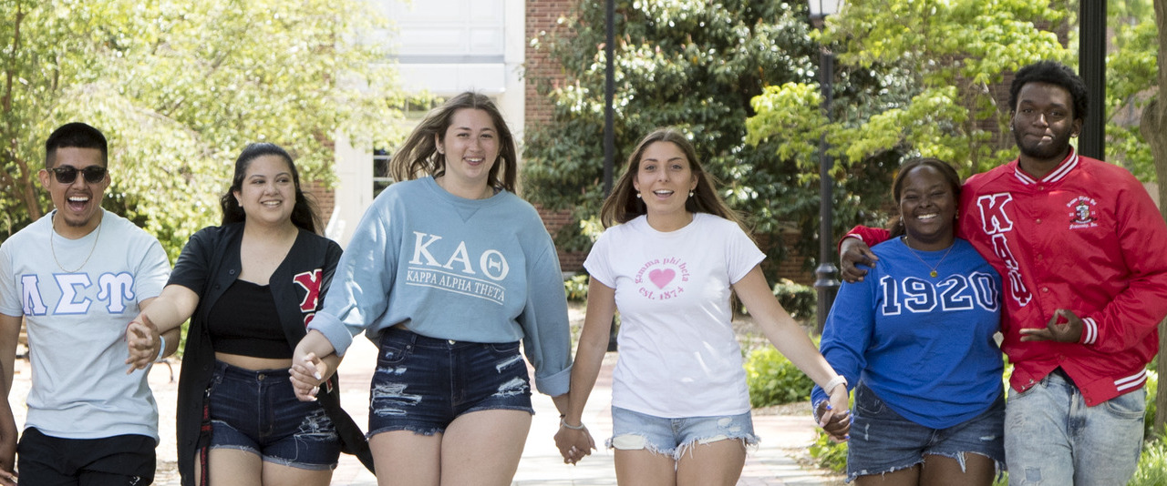 a group of students wearing fraternity and sorority shirts hold hands and walk toward the camera on the Green