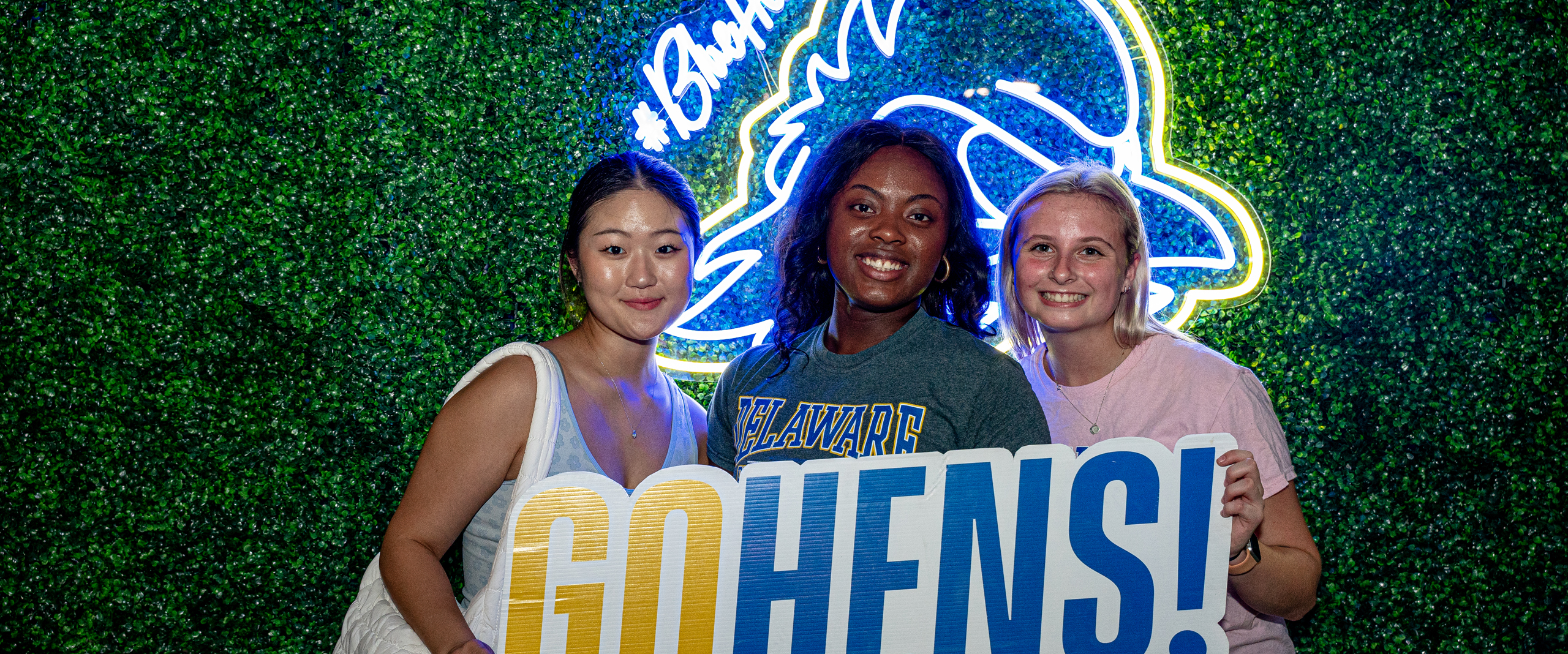 a trio of students poses in front of a photo backdrop holding a Go Hens sign