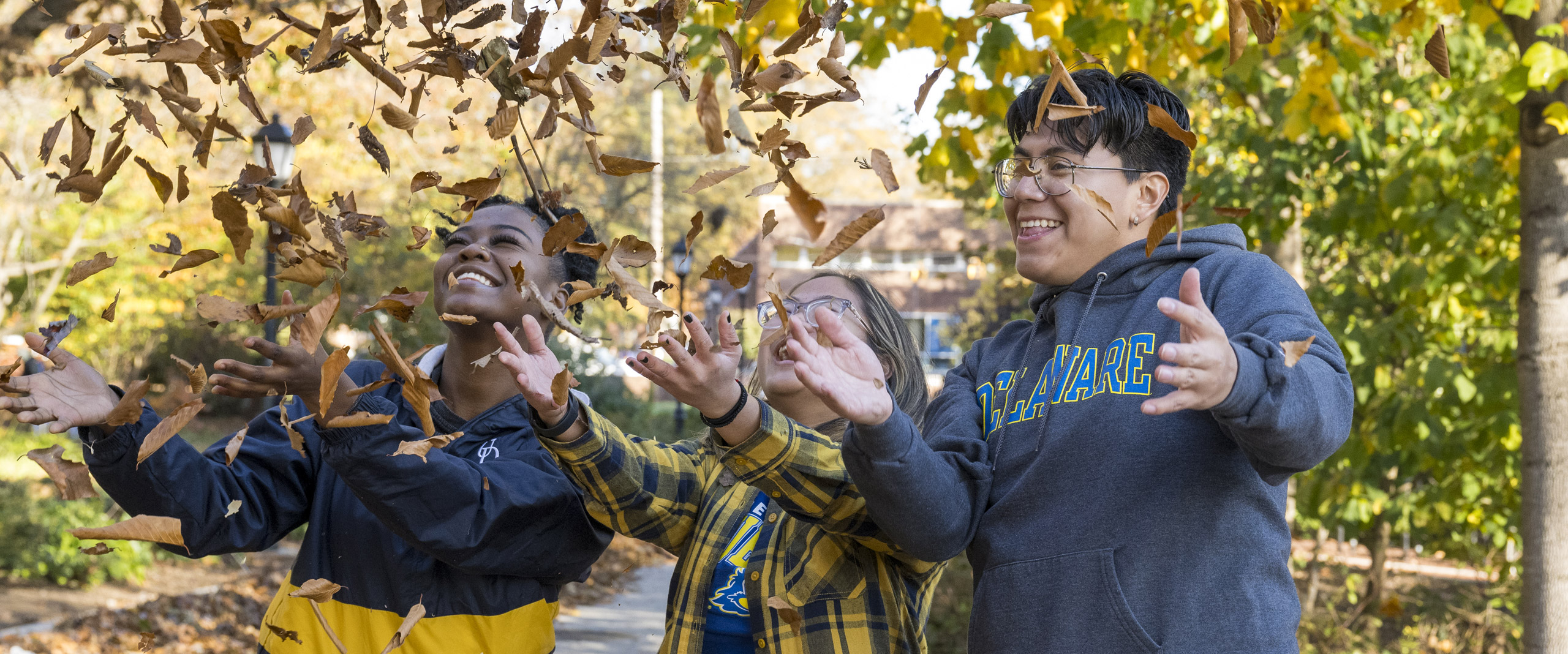 three students wear UD gear and celebrate fall by tossing leaves into the air while they laugh