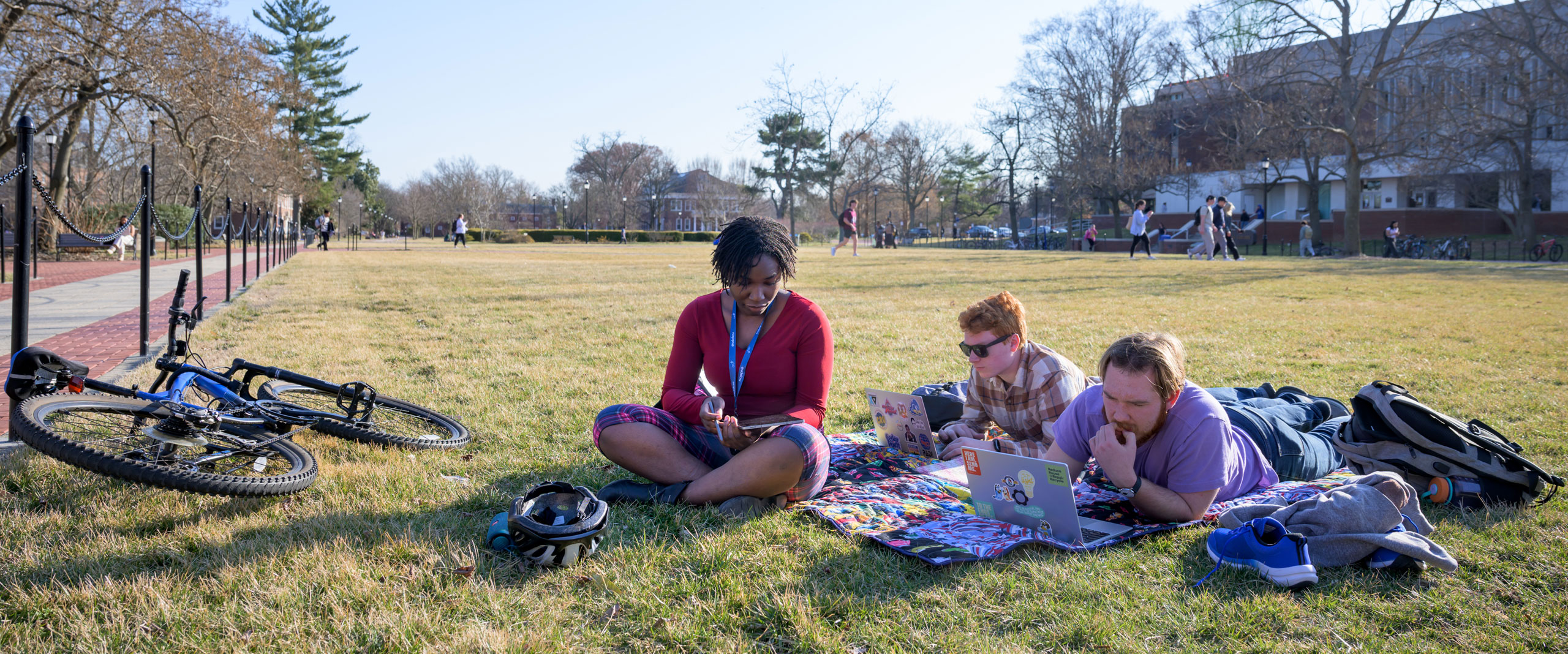 on a sunny day, three students sit and recline on The Green in front of University of Delaware's Morris Library.