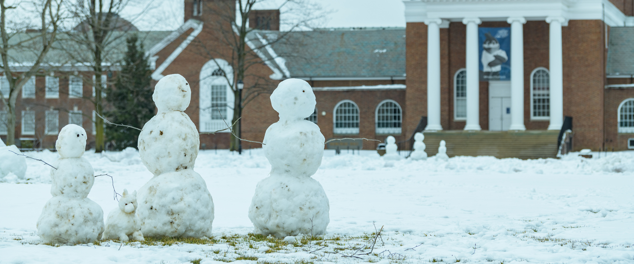 three student-built snowmen stand on University of Delaware's The Green with Memorial Hall in the background