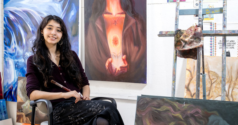 Isaura López, a senior fine arts major at the University of Delaware, in her studio in Taylor Hall. She imbues her paintings, like “¿Me Recuerdas?” and “Incubator” behind her, with physical makers of identity along with her personal relationships and heritage.