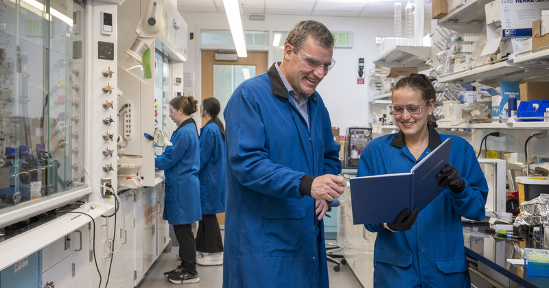 Two smiling researchers dressed in blue lab coats stand inside a university laboratory and look at a blue bound book of lab notes.