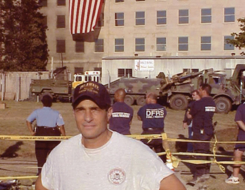 Man wearing UD cap stands in front of damaged Pentagon, a flag draped over its side.